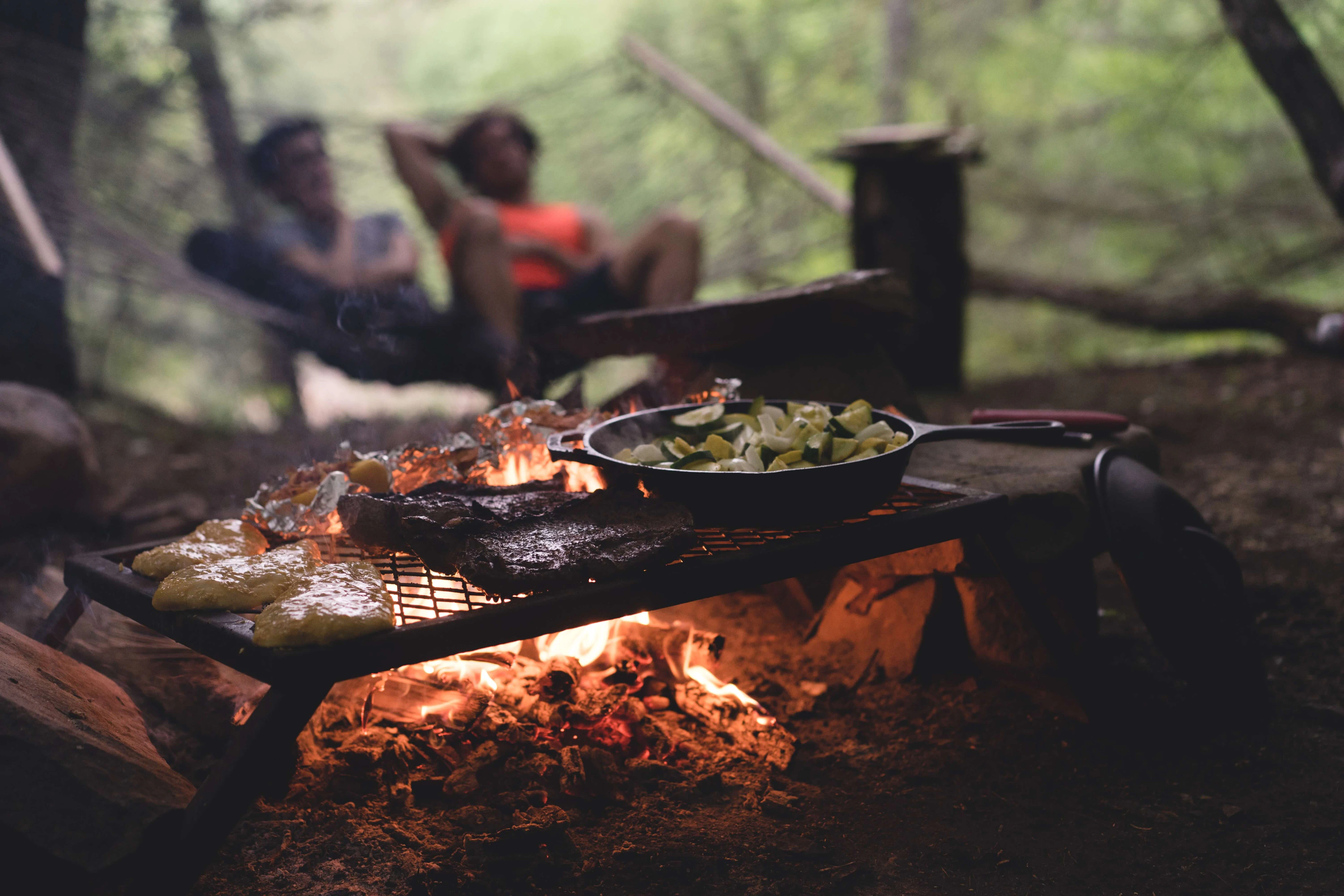 Food cooking over a campfire, with two friends blurred out in the background